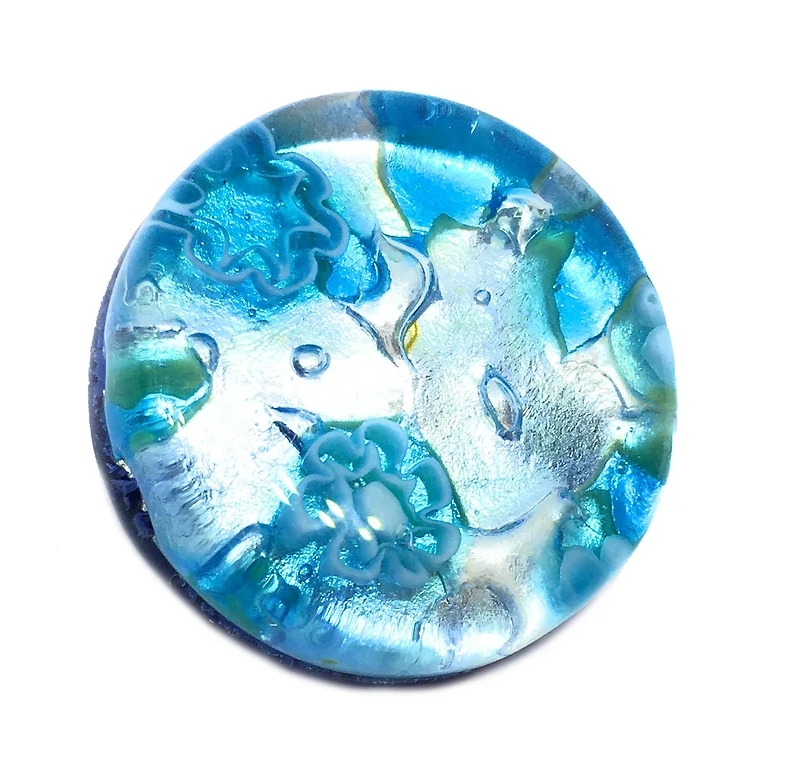 A Murano Passion brooch, measuring 2.4 cm in diameter and crafted with a silver leaf base, features a round design in translucent blue reminiscent of Venetian artistry. It showcases intricately carved fish and floral patterns, with its glossy finish and iridescent color variations enhancing its reflection of the rich heritage of Murano glasswork.