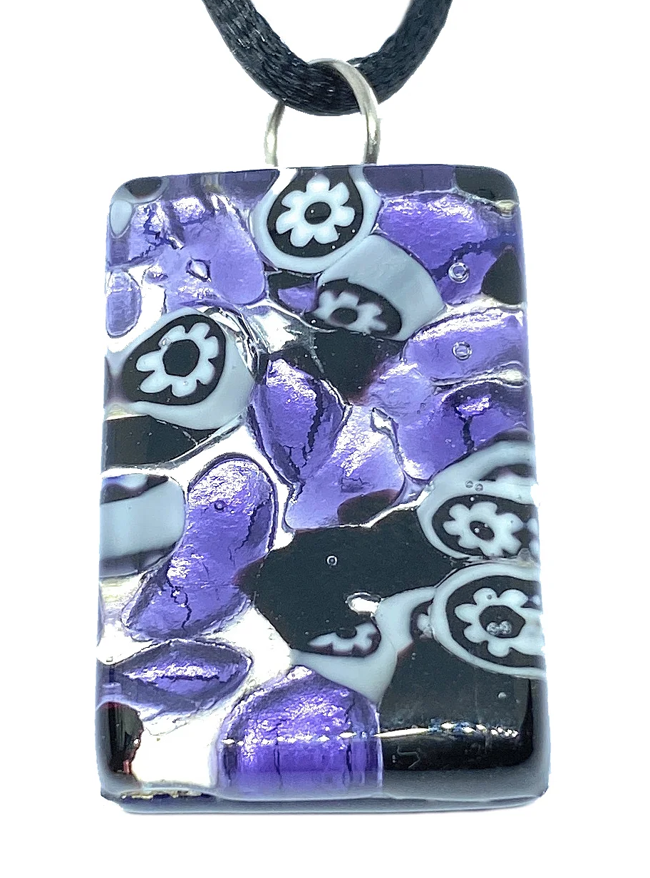 The Murano Passion pendant, crafted from genuine Millefiori Murano Glass, is a rectangular piece with dimensions of 3cm x 2cm. It hangs from a black cord and showcases an artistic mosaic design in shades of purple, black, and white. The pendant's intricate pattern combines floral motifs and abstract shapes against a silver leaf backdrop.