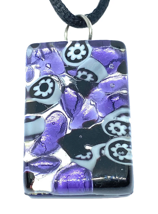 The Murano Passion pendant, crafted from genuine Millefiori Murano Glass, is a rectangular piece with dimensions of 3cm x 2cm. It hangs from a black cord and showcases an artistic mosaic design in shades of purple, black, and white. The pendant's intricate pattern combines floral motifs and abstract shapes against a silver leaf backdrop.