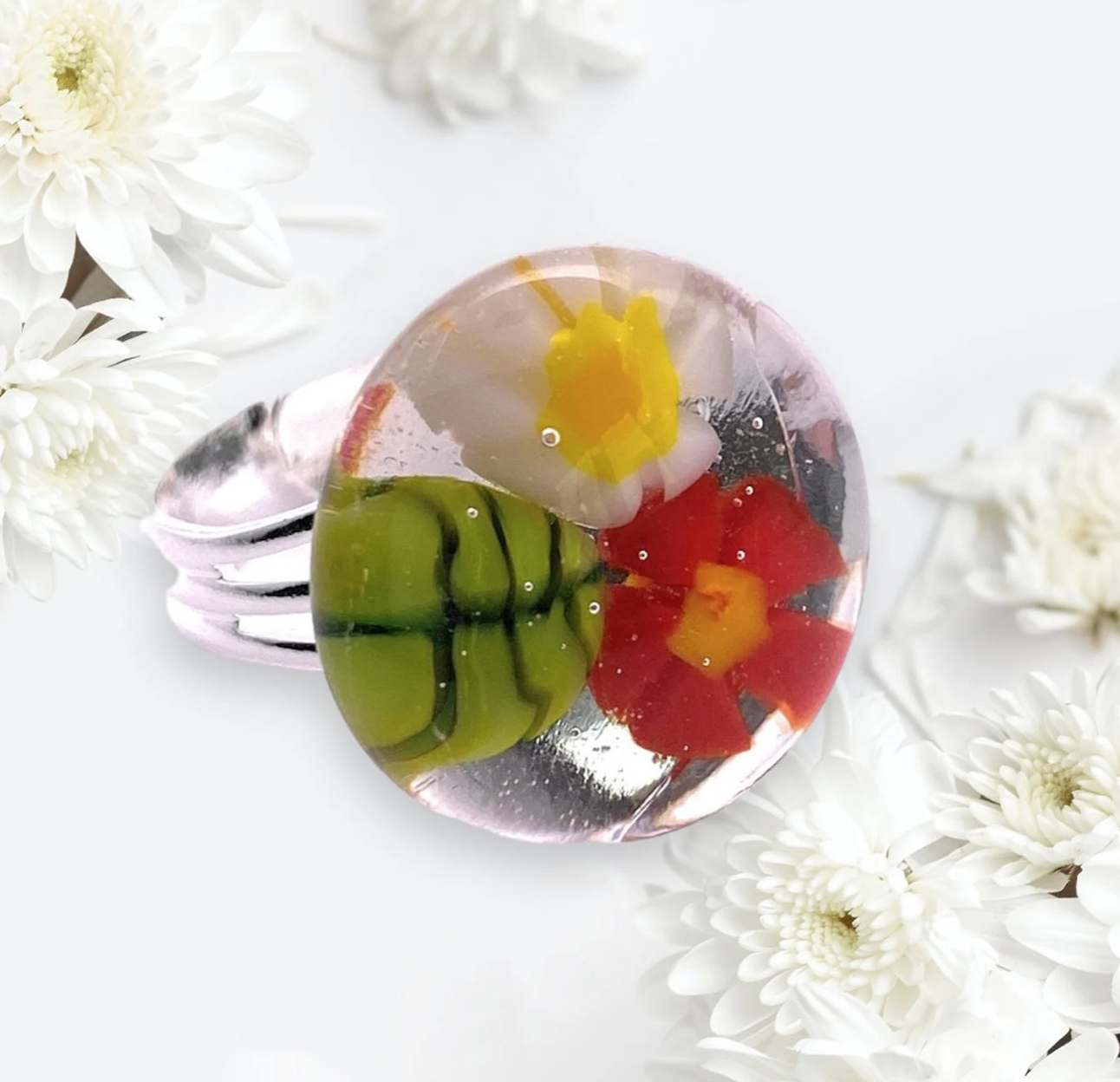 The Flower Ring by Calon Glass, also known as the Millefiori Ring or Handmade Glass Ring (CGR1), features a silver band topped with clear resin showcasing embedded red and yellow flowers and green leaves. The design is accented with white blooms on a light background, creating a stunning handmade glass effect.