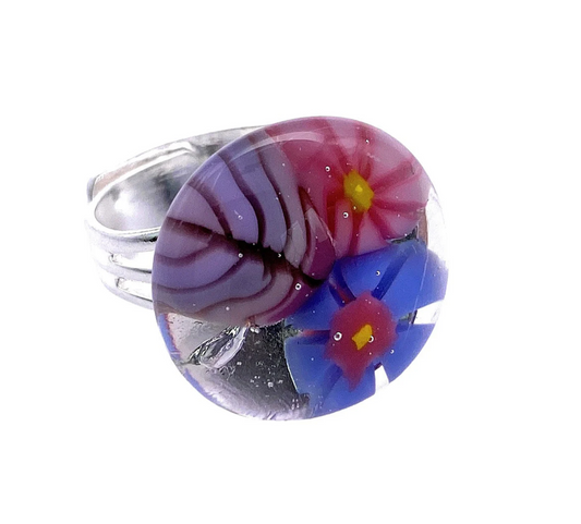 The Flower Ring by Calon Glass, also known as the Millefiori Ring (CGR2), is a silver ring that features a round handmade glass cabochon. It showcases an intricate Millefiori design with a pink flower that has a yellow center and a blue flower with a white center on its surface.