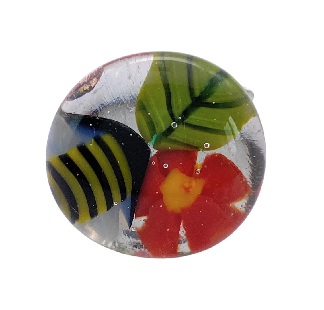 The Flower Ring, Millefiori Ring by Calon Glass (CGR3), is a captivating handmade glass ring featuring a vibrant and colorful design. It showcases an intricately crafted red flower with a yellow center and green leaves, complete with a bee-like striped element. The overall millefiori flower motif creates an artistic and bright effect.