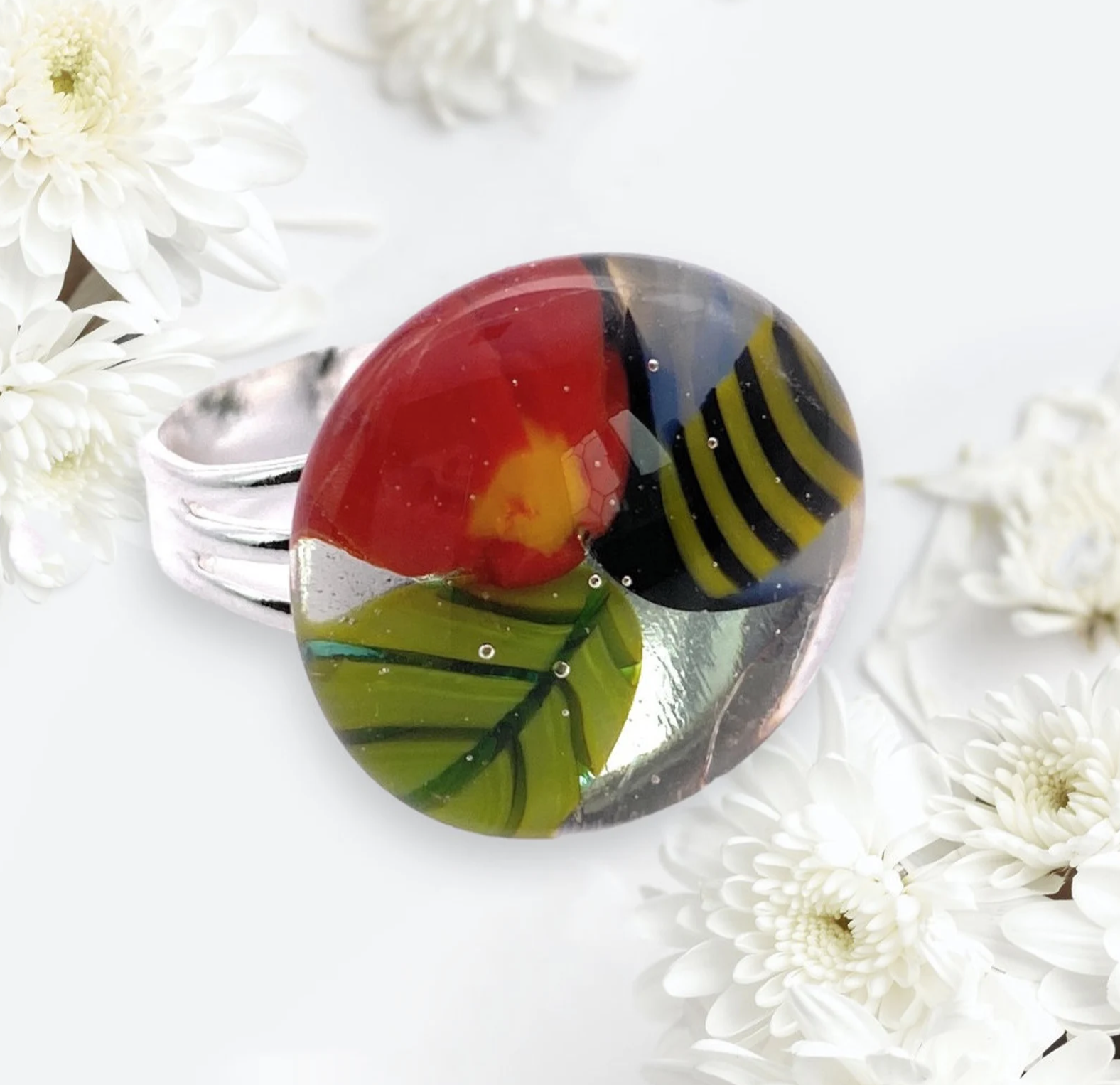 The Calon Glass Millefiori Ring, named CGR3, features a stunning array of abstract red, yellow, green, and black patterns set in silver. This handmade glass ring is beautifully complemented by white chrysanthemum flowers on a light background.