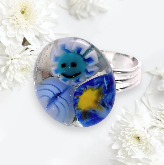 The Calon Glass Flower Ring, Millefiori Ring (CGR6), is a silver piece showcasing a lively, handmade glass design with a smiling sun and abstract patterns in blue and yellow. It is beautifully accentuated by soft-focus white chrysanthemums set against a light background.