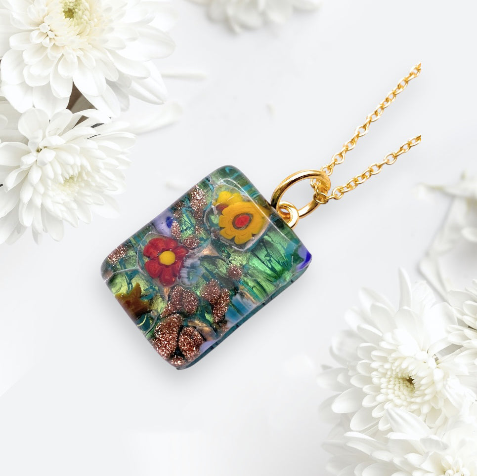 A vibrant Murano Passion Millefiori Flower Pendant on Gold Leaf, measuring a dainty 2cm x 1.5cm, hangs from a gold chain amidst white chrysanthemums on a light background. This exquisite piece of Murano Glass Jewellery guarantees elegance and authenticity.