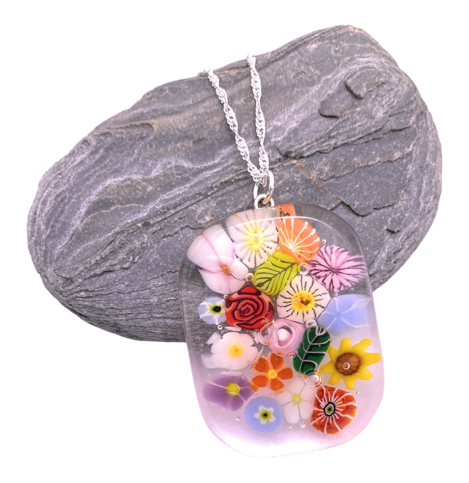 The Calon Glass Handmade Glass Pendant (CGP40) is a chunky 4cm x 3cm millefiori flower pendant on a silver chain, shown draped over a smooth, dark gray stone.