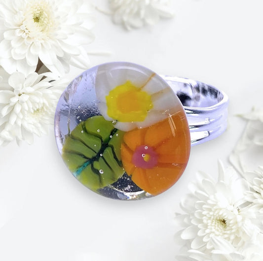 A Calon Glass masterpiece, the Flower Ring (CGR5) is a handmade Millefiori design with a striking resin dome featuring embedded colorful floral patterns in green, orange, and yellow. The silver ring is artistically displayed amidst delicate white chrysanthemum flowers on a light background.