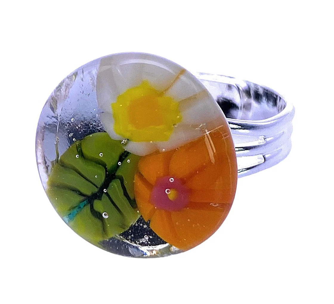 The Flower Ring by Calon Glass, a Millefiori ring known as CGR5, features a silver band with a round glass top. It displays three stunning hand-crafted flowers: one white with a yellow center, an orange flower with a pink center, and a green flower with a yellow center.