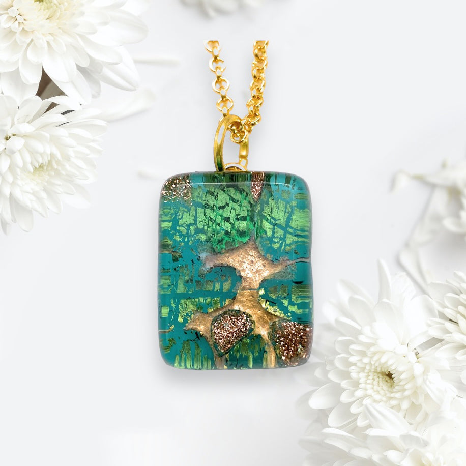 The Murano Passion Murano Glass Pendant, Green on Gold Leaf, measuring a dainty 2cm x 1.5cm (MGPP6), features an abstract pattern reminiscent of a tree against a light background. Accompanied by a gold chain and beautifully complemented by the delicate presence of white chrysanthemums, this exquisite piece of jewellery includes a Certificate of Authenticity.