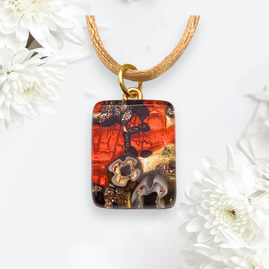 A lively Murano Glass Pendant from Murano Passion, showcasing a fusion of red, orange, and brown tones with abstract designs and a glossy finish, elegantly dangles from a brown cord. Set against a light backdrop adorned with white flowers on the sides, this exquisite Millefiori Pendant on Gold Leaf (MGPP26) measuring 2cm x 1.5cm includes a Certificate of Authenticity.