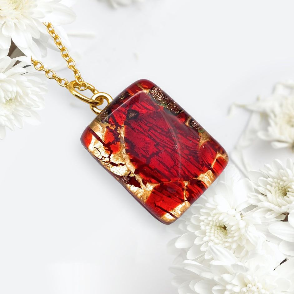 A Murano Passion creation, the Murano Glass Pendant on Gold Leaf (MGPP24) showcases a dainty 2cm x 1.5cm pendant with exquisite red and gold marbling, gracefully suspended from a gold chain. This remarkable piece is beautifully highlighted against delicate white flowers on a light background and includes a Certificate of Authenticity to affirm its elegance and craftsmanship.